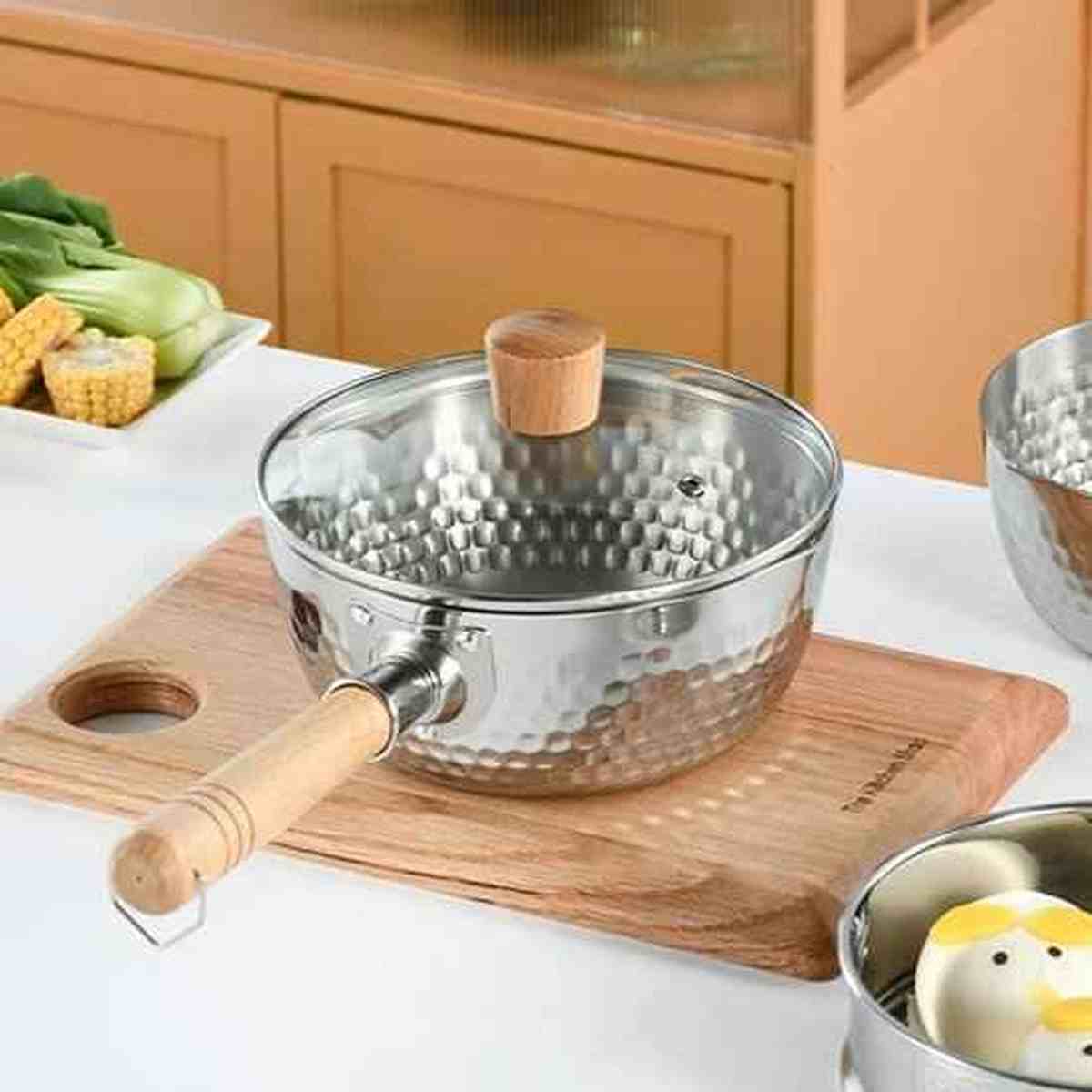 Casserole en acier inoxydable martelé avec son couvercle en verre trempé et pommeau en bois assorti sur une planche à découper.
