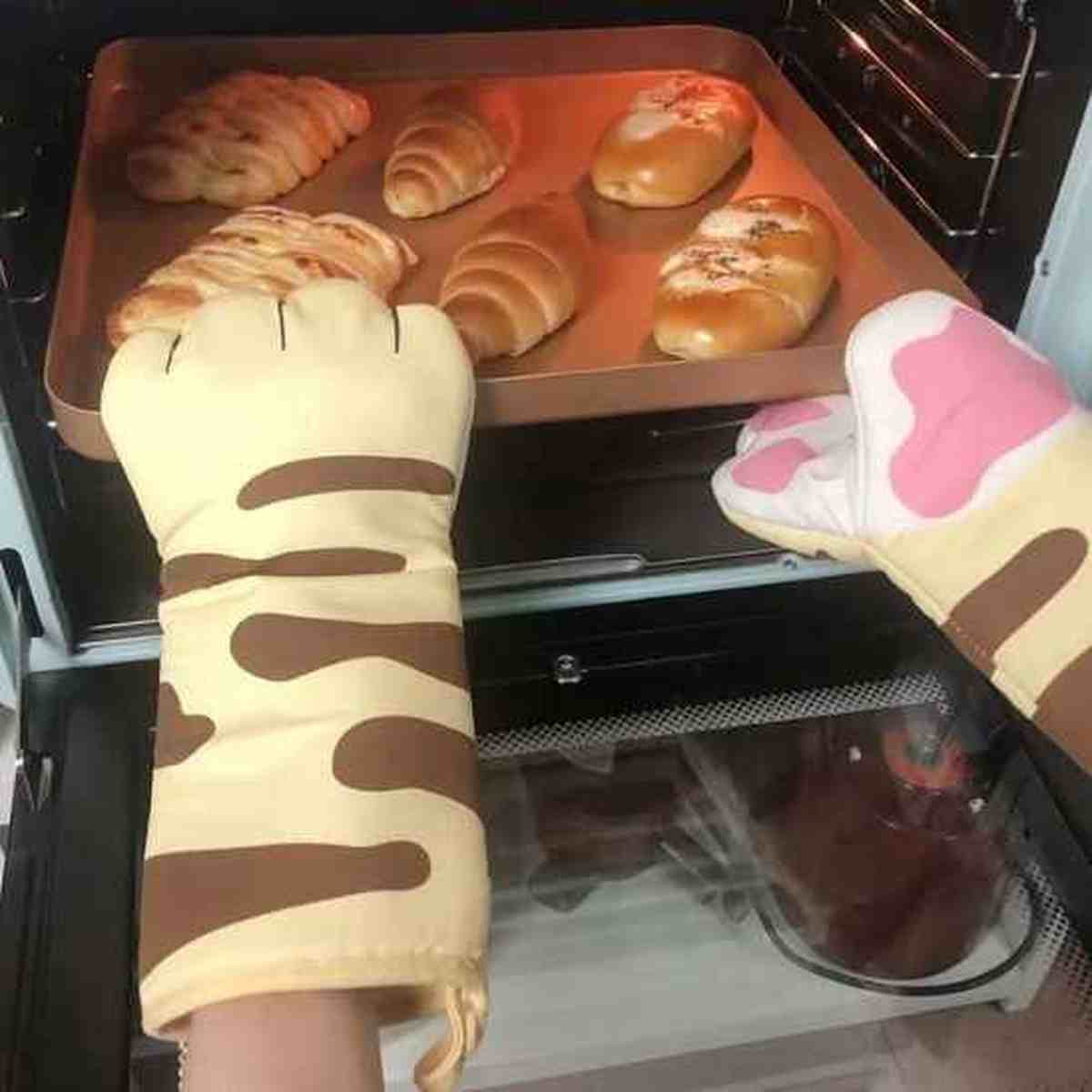 Paire de moufles de cuisine beiges avec motifs pattes de chat sortant une plaque de croissants chauds du four, protection 300°C.