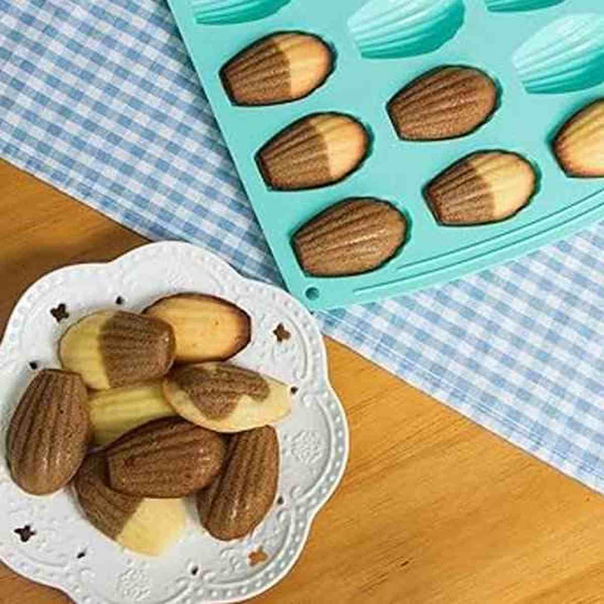 Moule à madeleines en silicone bleu ciel PâtissiPro avec des madeleines bicolores nature et chocolat, posé sur une nappe à carreaux bleue.