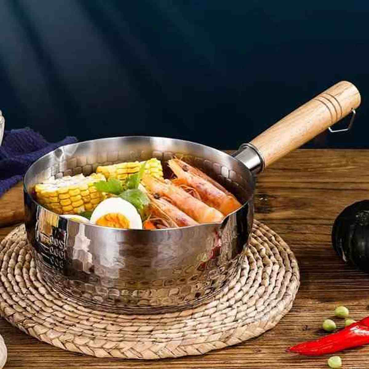 Casserole design en acier inoxydable martelé avec manche en bois, contenant un bouillon de crevettes, maïs et œufs sur un dessous de plat.