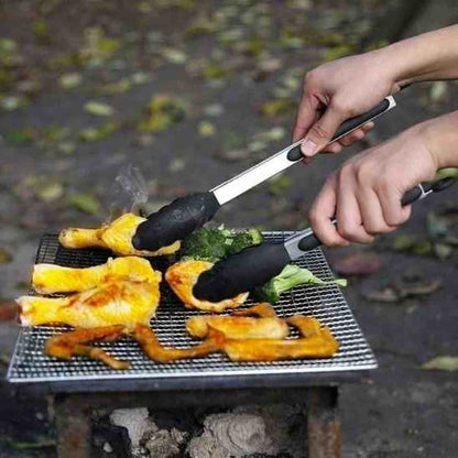 Pince à barbecue en acier inoxydable et silicone noir utilisée pour retourner du poulet et des brocolis sur une grille de barbecue en extérieur.