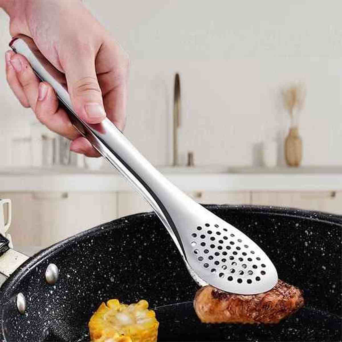 Pince de cuisine en acier inoxydable avec tête perforée saisissant de la viande et du maïs dans une poêle, illustrant le contrôle et la précision.