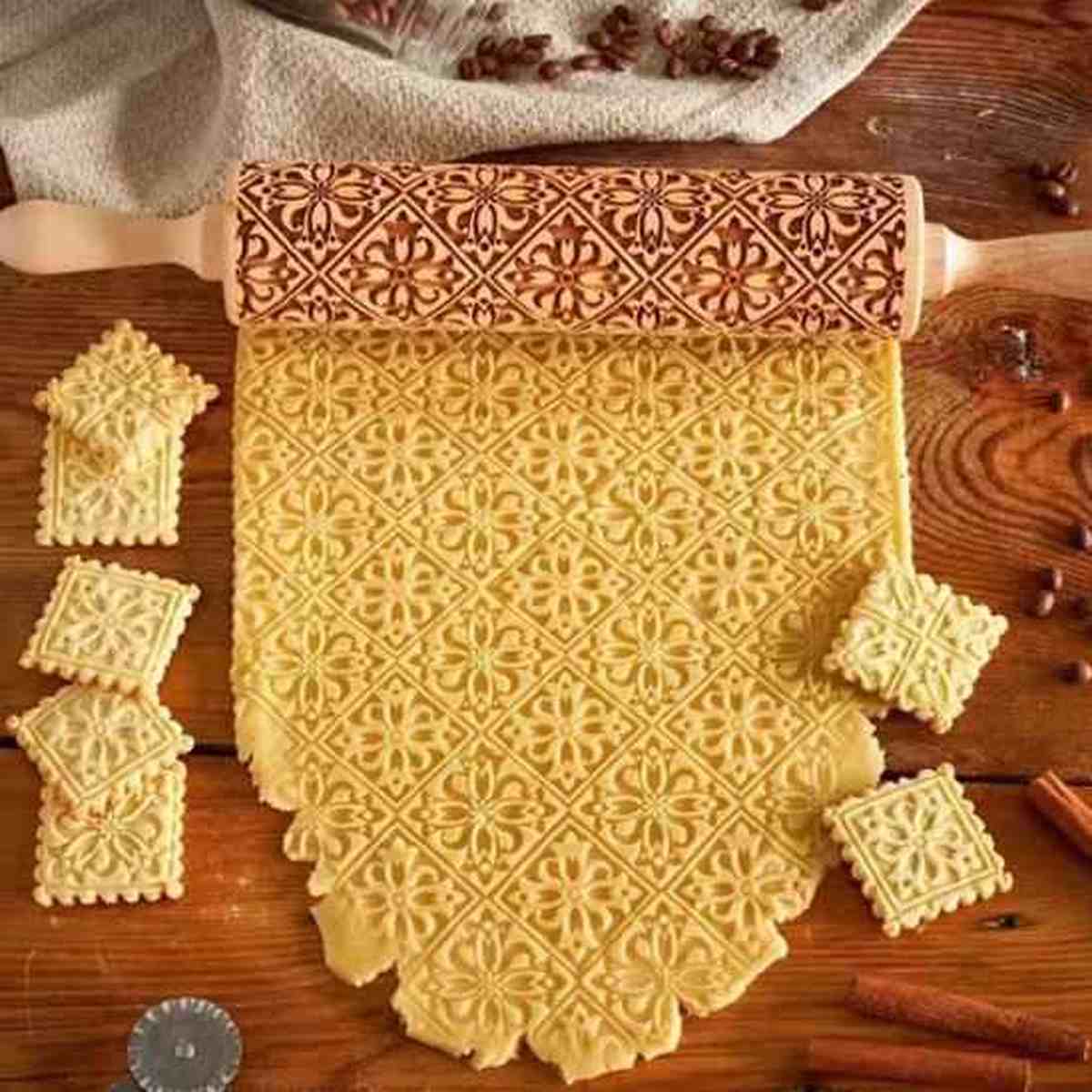 Rouleau à pâtisserie en bois gravé posé sur une pâte à biscuits affichant des motifs floraux et géométriques détaillés, avec des biscuits découpés à côté.