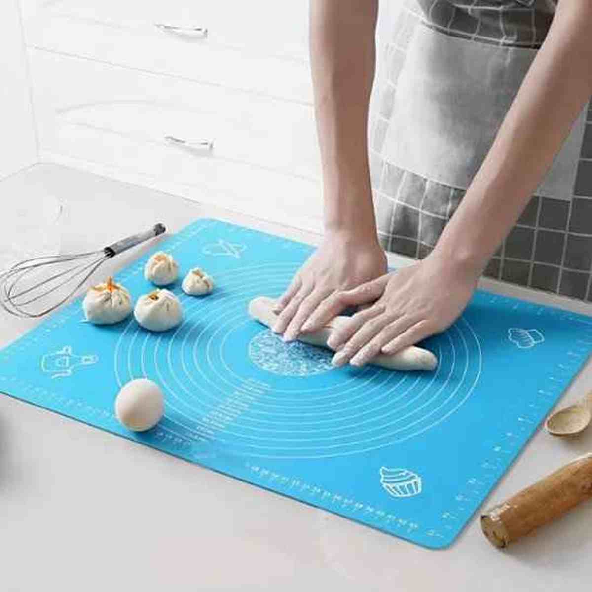 Mains pétrissant de la pâte sur un tapis de pâtisserie bleu gradué, entouré de petits pains formés et d'un fouet, illustrant la stabilité du silicone